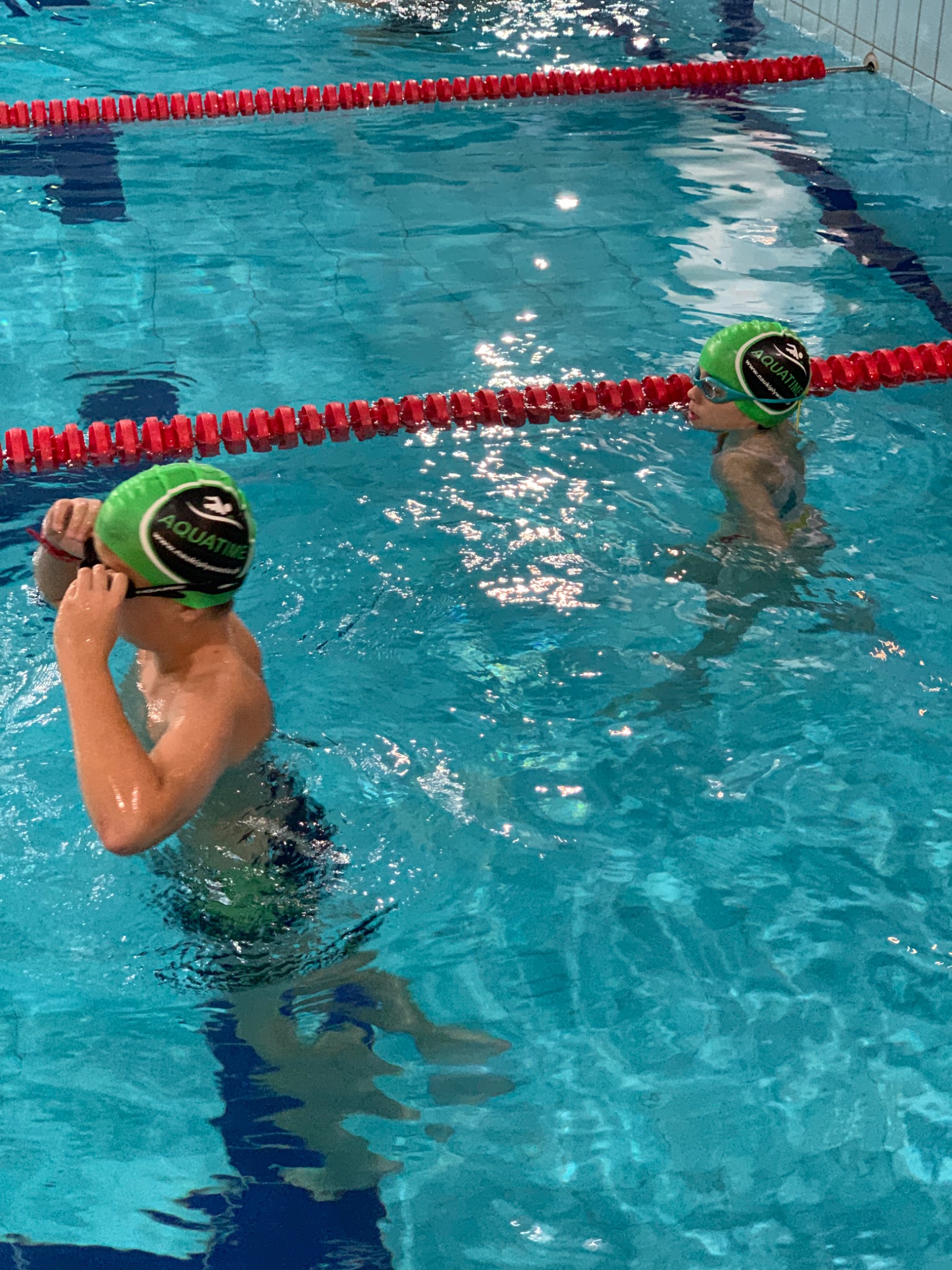 Dwoje dzieci w zielonych czepkach i okularach pływackich w basenie z błękitną wodą, oddzielonym czerwonymi linami.