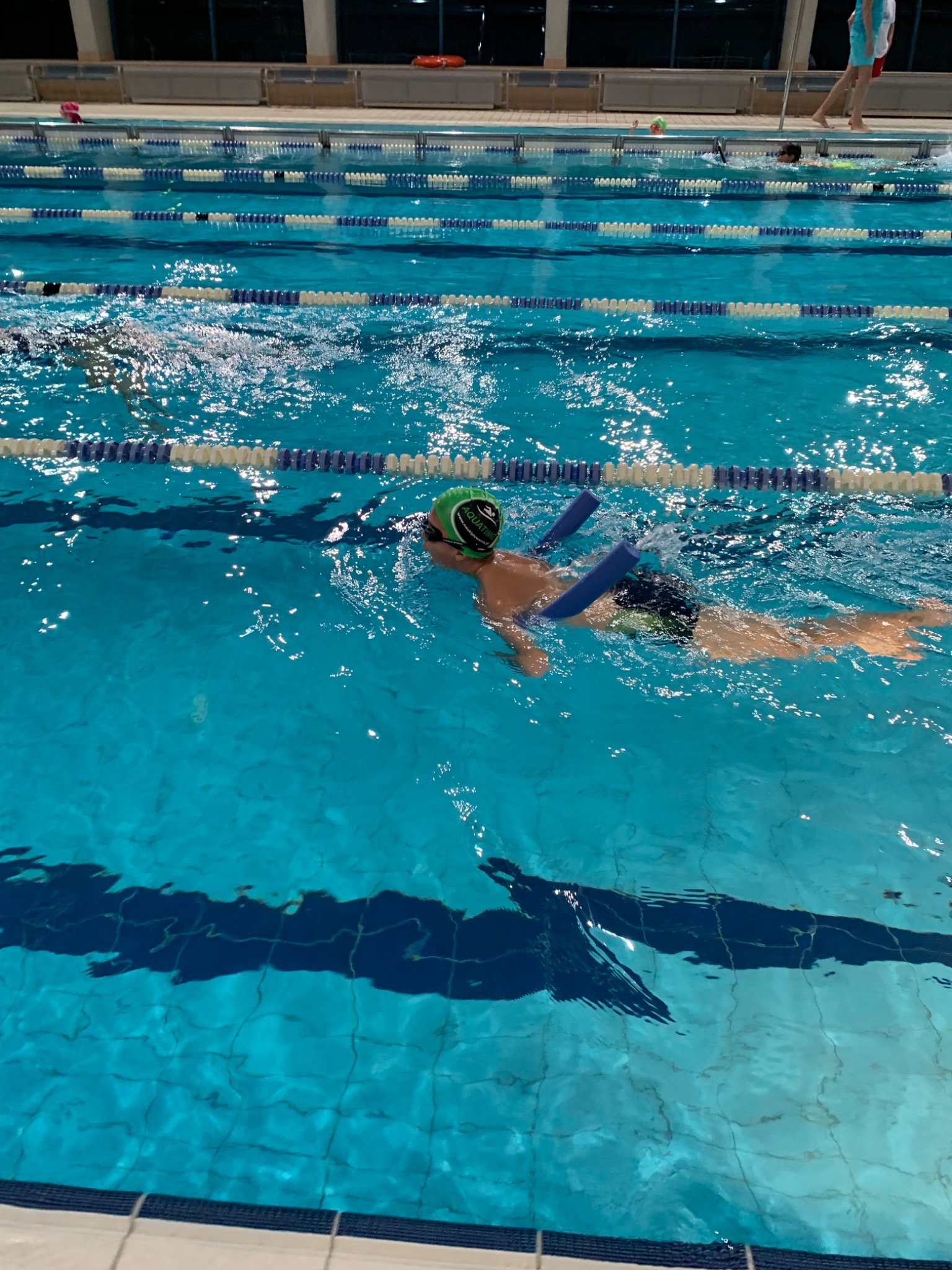 Chłopiec w czepku i okularach pływackich, z piankowymi rurkami pod pachami, unosi się na wodzie w basenie z wyznaczonymi torami.