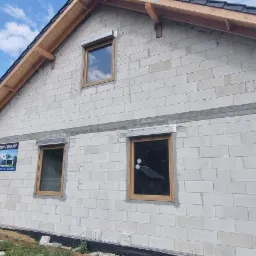 Ściana domu w trakcie budowy z zamontowanymi oknami w okleinie Woodeck Oak, widoczny plakat reklamowy firmy Fidia okna/drzwi/bramy.