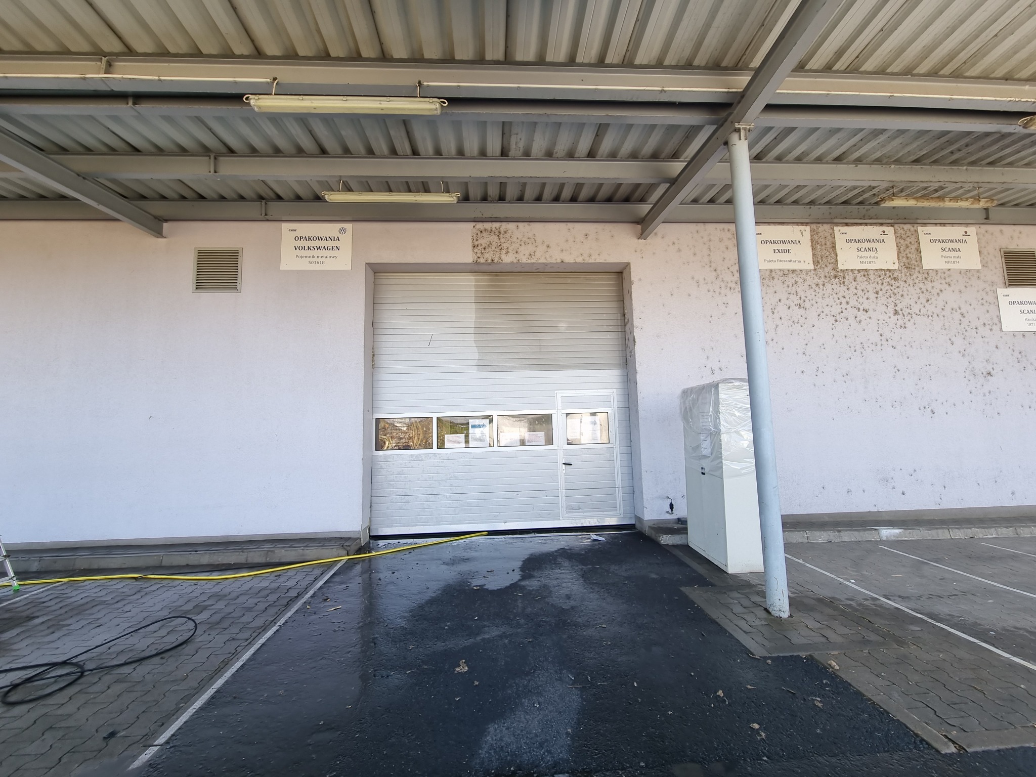 Biała elewacja budynku magazynowego z bramą rolowaną, widoczne ślady zabrudzeń i wilgoci po czyszczeniu, kostka brukowa mokra, żółty wąż ogrodowy leżący na ziemi.