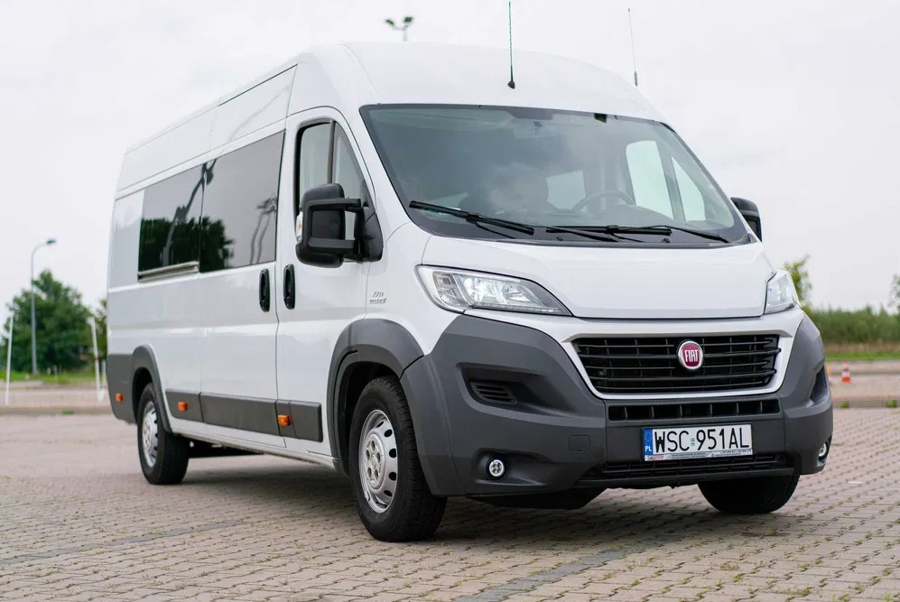 Biały, wysoki Fiat Ducato z przyciemnianymi szybami, zaparkowany na brukowanym placu, widok z przodu na tablicę rejestracyjną WSC 951AL.