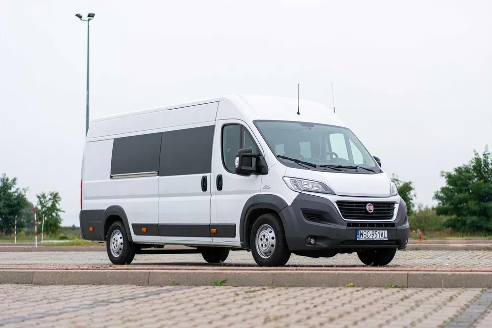 Biały bus Fiat Ducato z przyciemnianymi szybami na parkingu z kostki brukowej, widok z przodu i boku, tablica rejestracyjna WSC 951AL.