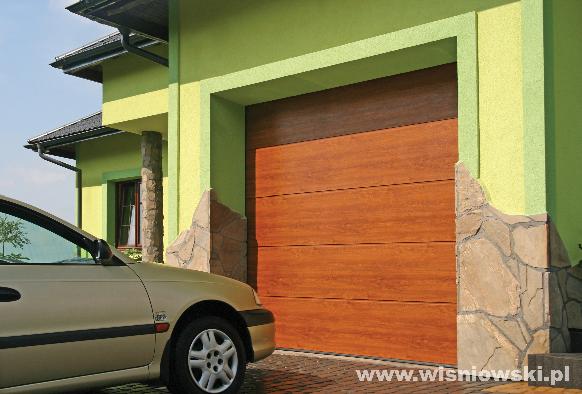 Brązowa segmentowa brama garażowa z imitacją drewna, zamontowana w zielonym budynku z kamienną elewacją, obok zaparkowany beżowy samochód.