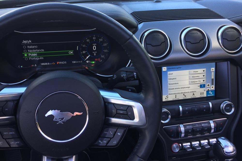 Wnętrze samochodu z widocznym kokpitem, kierownicą z logo konia, ekranem dotykowym ustawionym na język polski i panelem klimatyzacji.