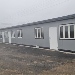 BPJA Paweł Bernacki - Szary budynek modułowy z białymi oknami i drzwiami, widok z zewnątrz na brukowanym placu, z widocznym wiadrem farby przy ścianie.