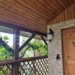 BPJA Paweł Bernacki - Zadaszony ganek z drewnianą boazerią sufitową, kamienną elewacją, drewnianymi drzwiami z ozdobnym wieńcem i kutą lampą ścienną w kształcie konia