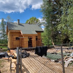 BPJA Paweł Bernacki - Wznoszony dom szkieletowy z zielonym dachem, widok przez ogrodzenie z siatki, rusztowania przy ścianach, pracownik na dachu, w tle drzewa i błękitne niebo z chmurami.