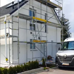 Dwukondygnacyjny dom z białymi ścianami i ciemnym dachem, otoczony rusztowaniem, z widocznym fragmentem białego samochodu dostawczego z przodu.