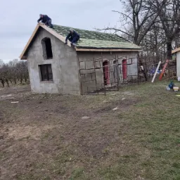 Dwóch dekarzy montuje łatowanie na dachu surowego domu z pustaków, widoczne rusztowanie, okno i otwór drzwiowy bez futryn.