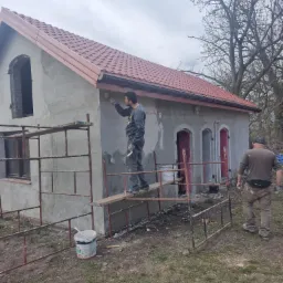 Pracownik na rusztowaniu maluje elewację budynku z pomalowanym na czerwono dachem, widoczny drugi pracownik i wiadro farby, szara, pochmurna pogoda.
