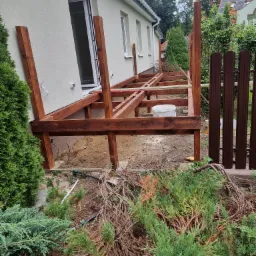 Konstrukcja drewnianego tarasu przy domu jednorodzinnym, widoczne słupy i belki nośne, białe okno balkonowe, elementy ogrodzenia w tle, etap budowy.