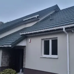 Jednopiętrowy dom jednorodzinny z ciemnozielonym dachem z blachodachówki, jasną elewacją i białymi rynnami, widziany z zewnątrz w pochmurny dzień.