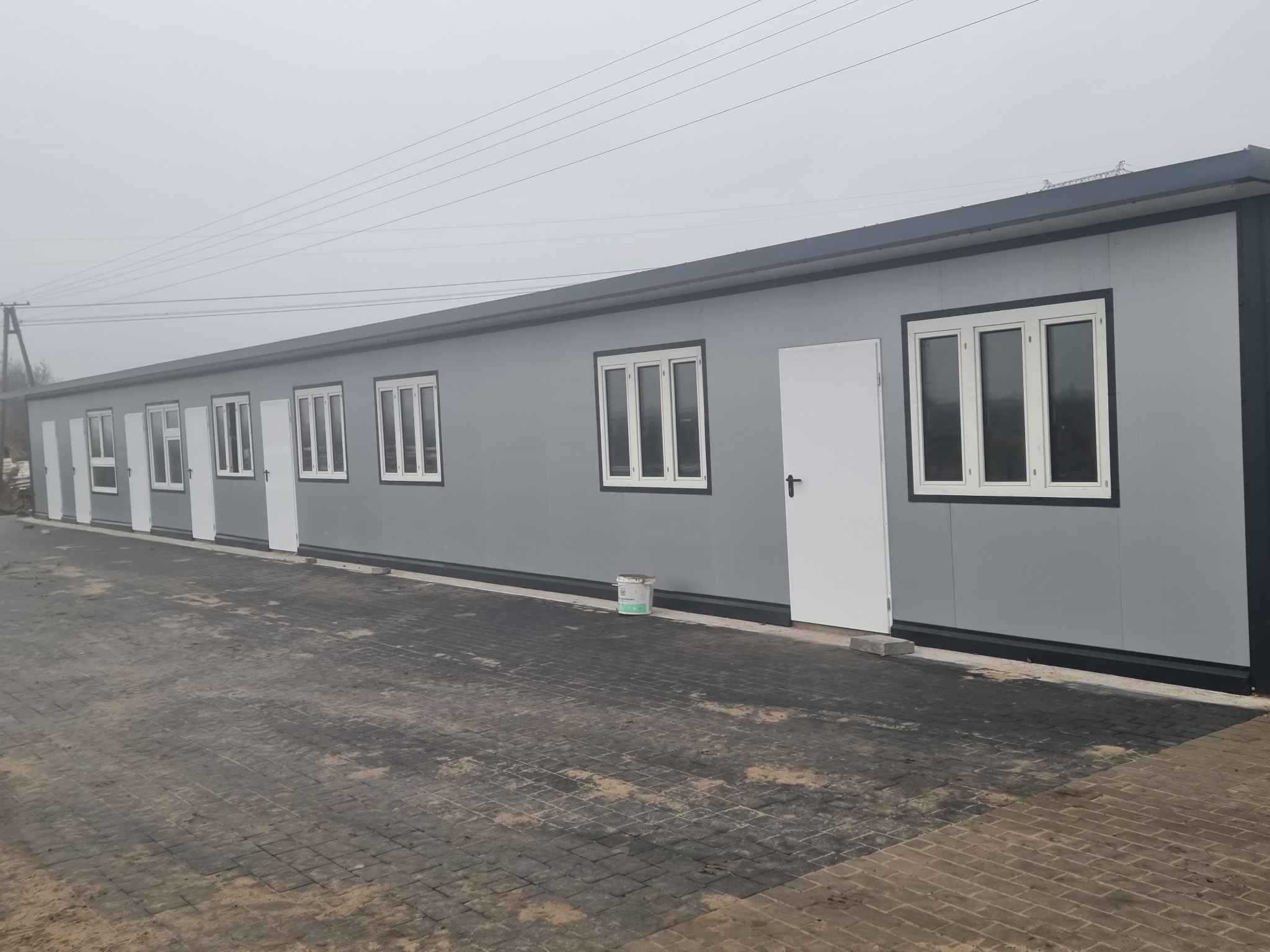 Szary budynek modułowy z białymi oknami i drzwiami, widok z zewnątrz na brukowanym placu, z widocznym wiadrem farby przy ścianie.