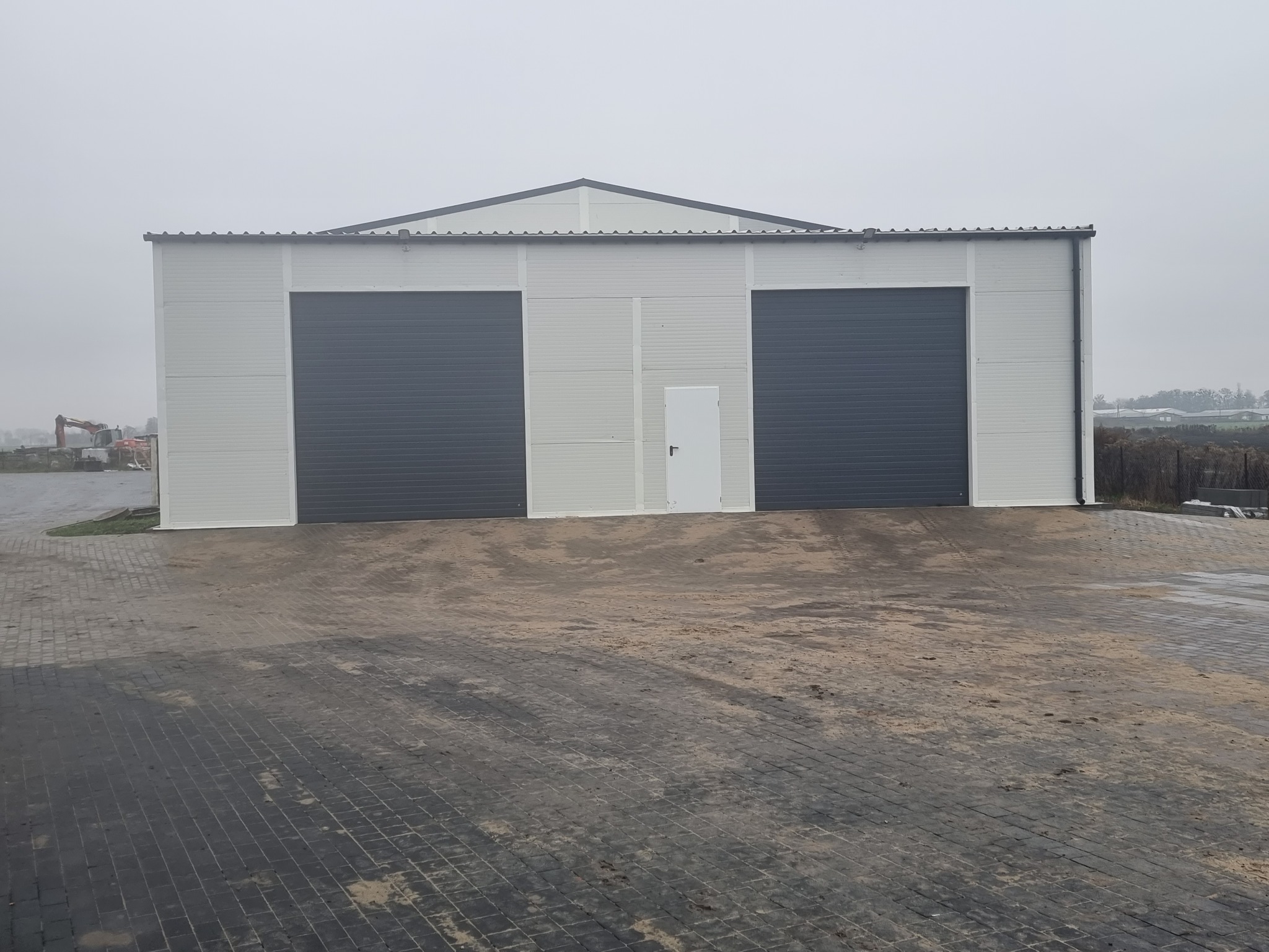 Nowoczesny budynek magazynowy z dwoma szarymi bramami segmentowymi i białymi panelami elewacyjnymi, widok zewnętrzny w pochmurny dzień.