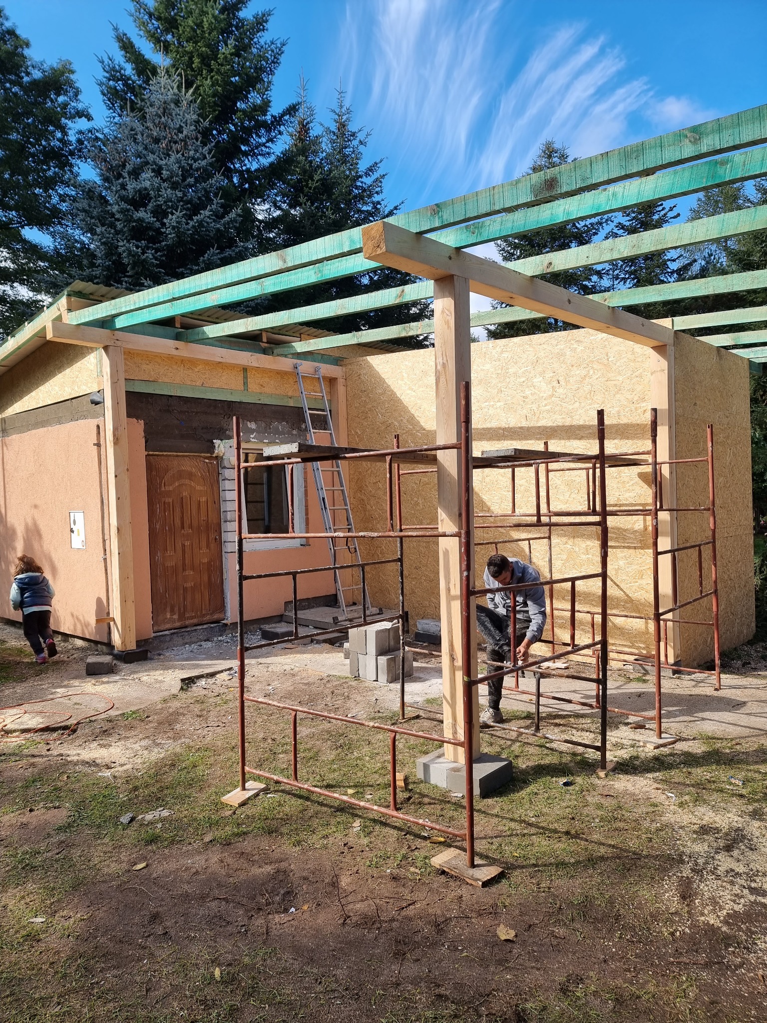 Budowa małego domu z drewnianą konstrukcją szkieletową i płyt OSB, widoczny pracownik na rusztowaniu oraz częściowo otynkowana ściana z brązowymi drzwiami.