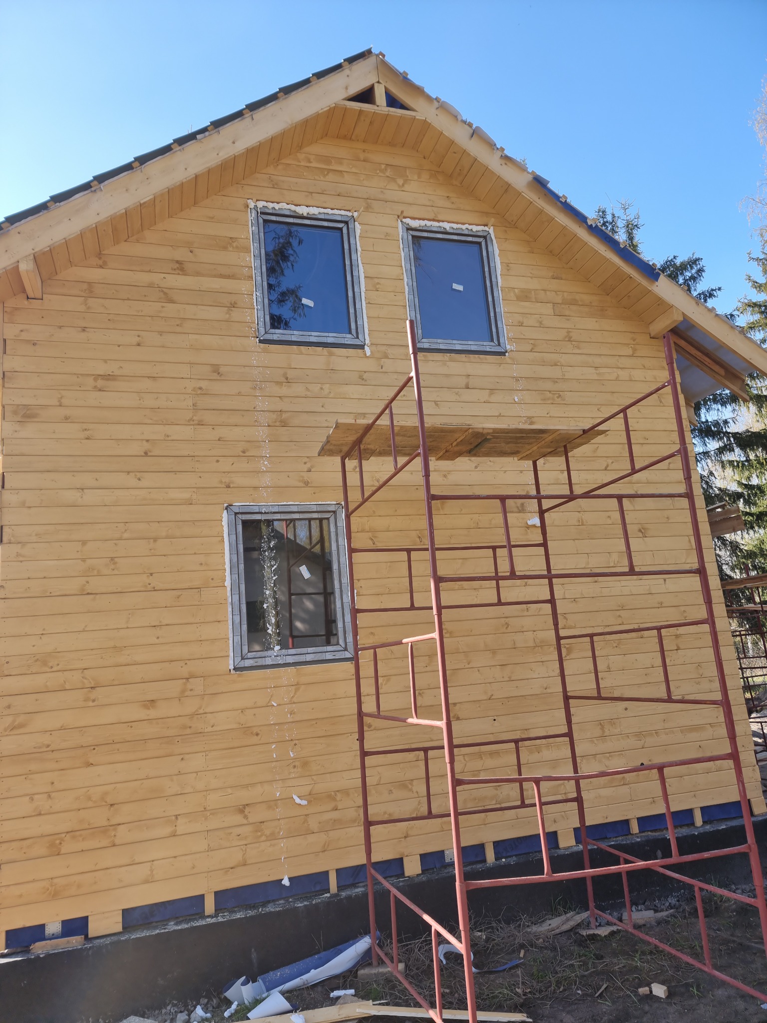 Wznoszony dom z jasnego drewna z widocznymi rusztowaniami, trzema oknami z folią ochronną i przygotowanym fundamentem, fotografowany w słoneczny dzień.