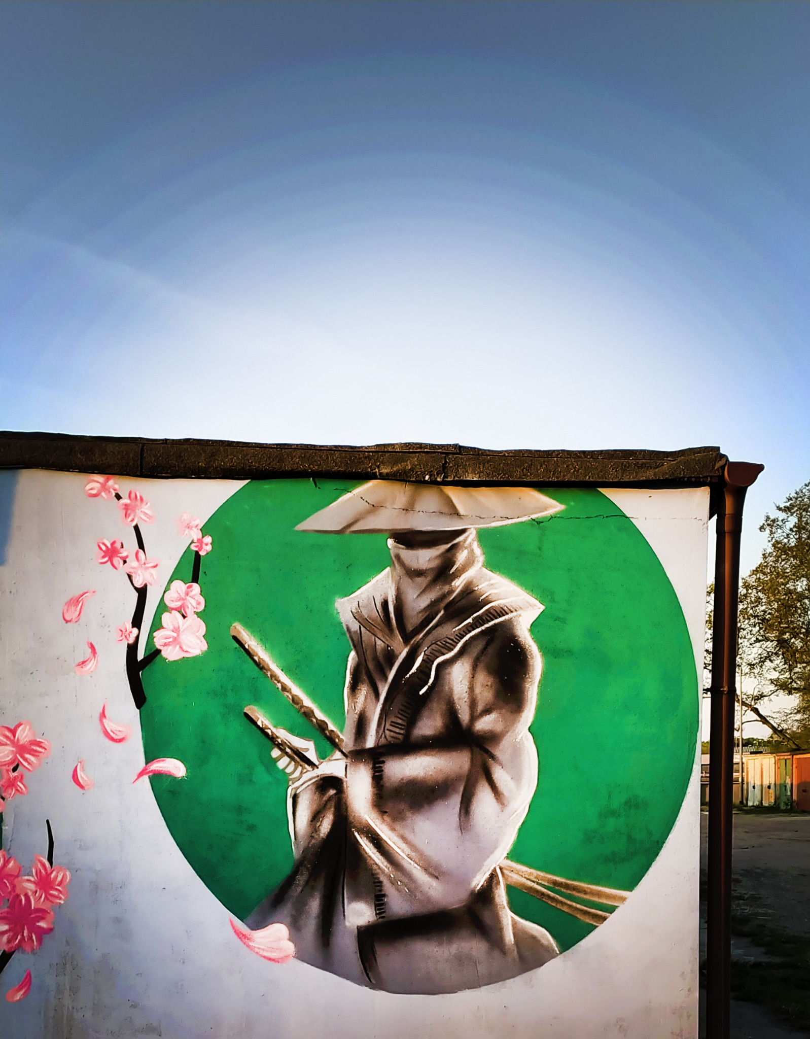 Graffiti przedstawiające samuraja w kapeluszu, z mieczami i kwitnącą wiśnią na ścianie budynku.