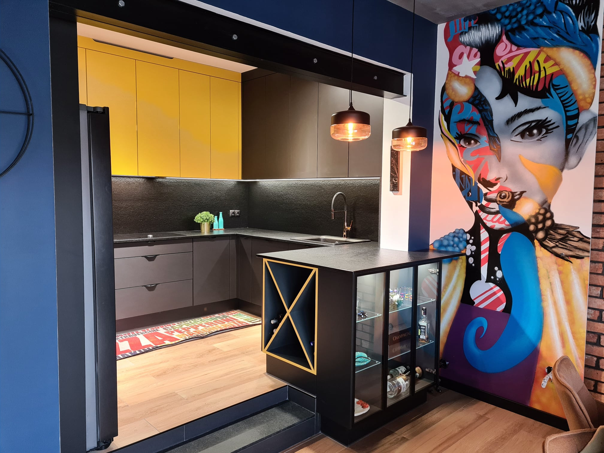 Nowoczesna kuchnia z żółtymi szafkami górnymi, czarnymi szafkami dolnymi i wyspą z ekspozycją na wino, uzupełniona o mural z portretem kobiety w stylu pop-art na ścianie.