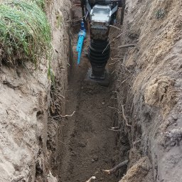 HYDROREM - Pracownik w pomarańczowej kamizelce zagęszcza ziemię w wykopie za pomocą zagęszczarki wibracyjnej typu skoczek; widoczne korzenie i gałęzie w ziemi.