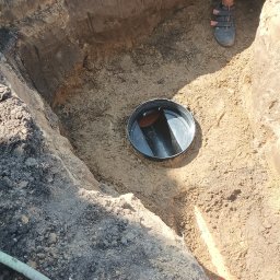 HYDROREM - Wykop w ziemi z widocznym czarnym zbiornikiem z rurami, prawdopodobnie na szambo lub drenaż, z fragmentami rur zielonej i pomarańczowej na krawędzi wykopu i fragmentem osoby w prawym górnym rogu.