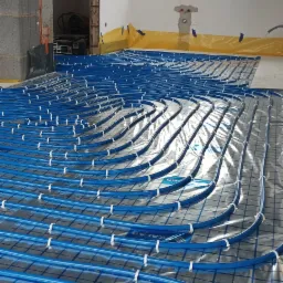 Niebieskie rury ogrzewania podłogowego ułożone na folii z siatką, przymocowane białymi klipsami, widoczne okno i surowy stan wykończenia pomieszczenia.