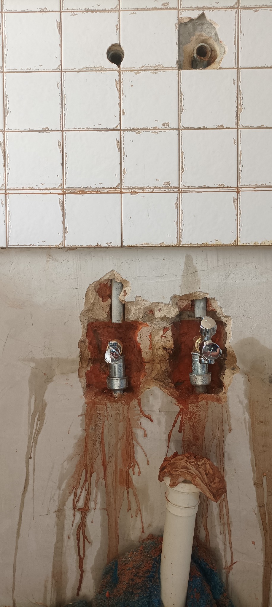 Odsłonięte rury z zaworami w ścianie łazienki podczas prac remontowych, widoczne fragmenty starej instalacji i uszkodzone płytki.