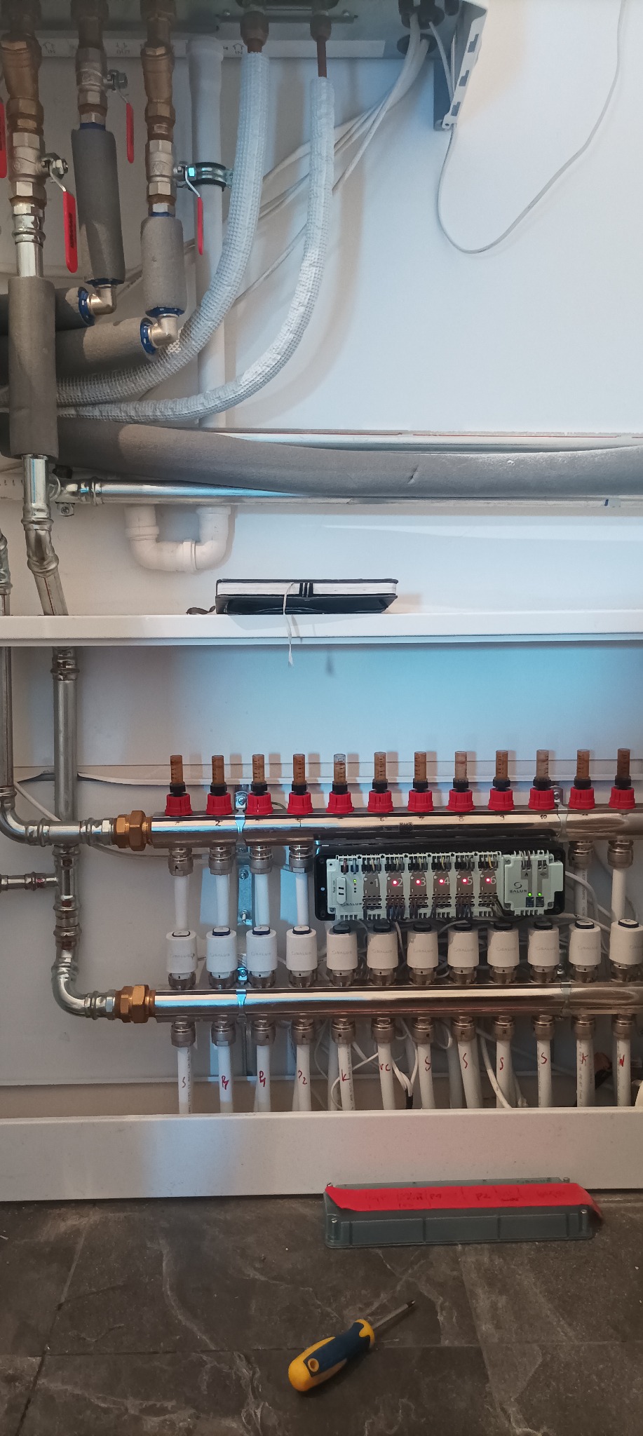 Rozdzielacz ogrzewania podłogowego z rurami, zaworami i sterownikiem elektronicznym Salus, widoczny również śrubokręt na podłodze z płytek imitujących marmur.