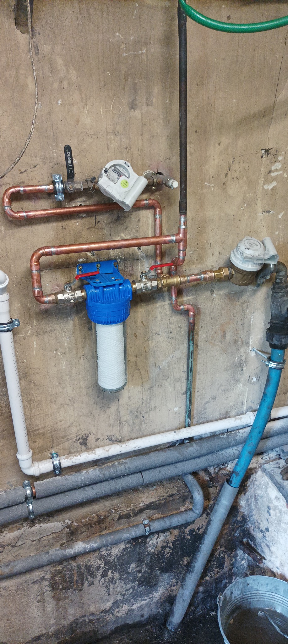 Filtr wody z niebieską obudową i wkładem sznurkowym, zamontowany na miedzianych rurach w pomieszczeniu z surowymi ścianami i widocznymi instalacjami.