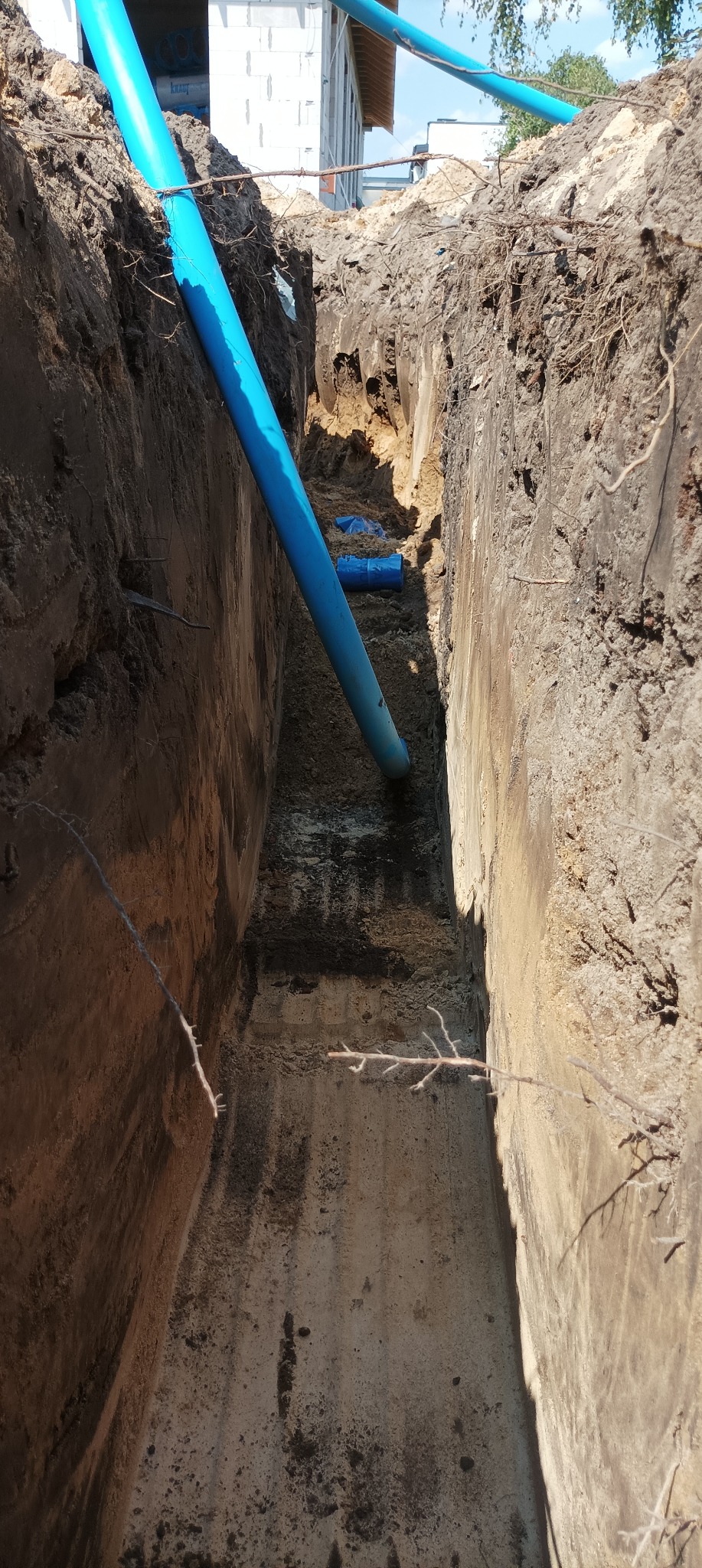 Niebieska rura wodociągowa ułożona ukośnie w wykopie ziemnym, widok z góry na dno wykopu z fragmentami innych rur. W tle widoczny budynek w trakcie budowy.