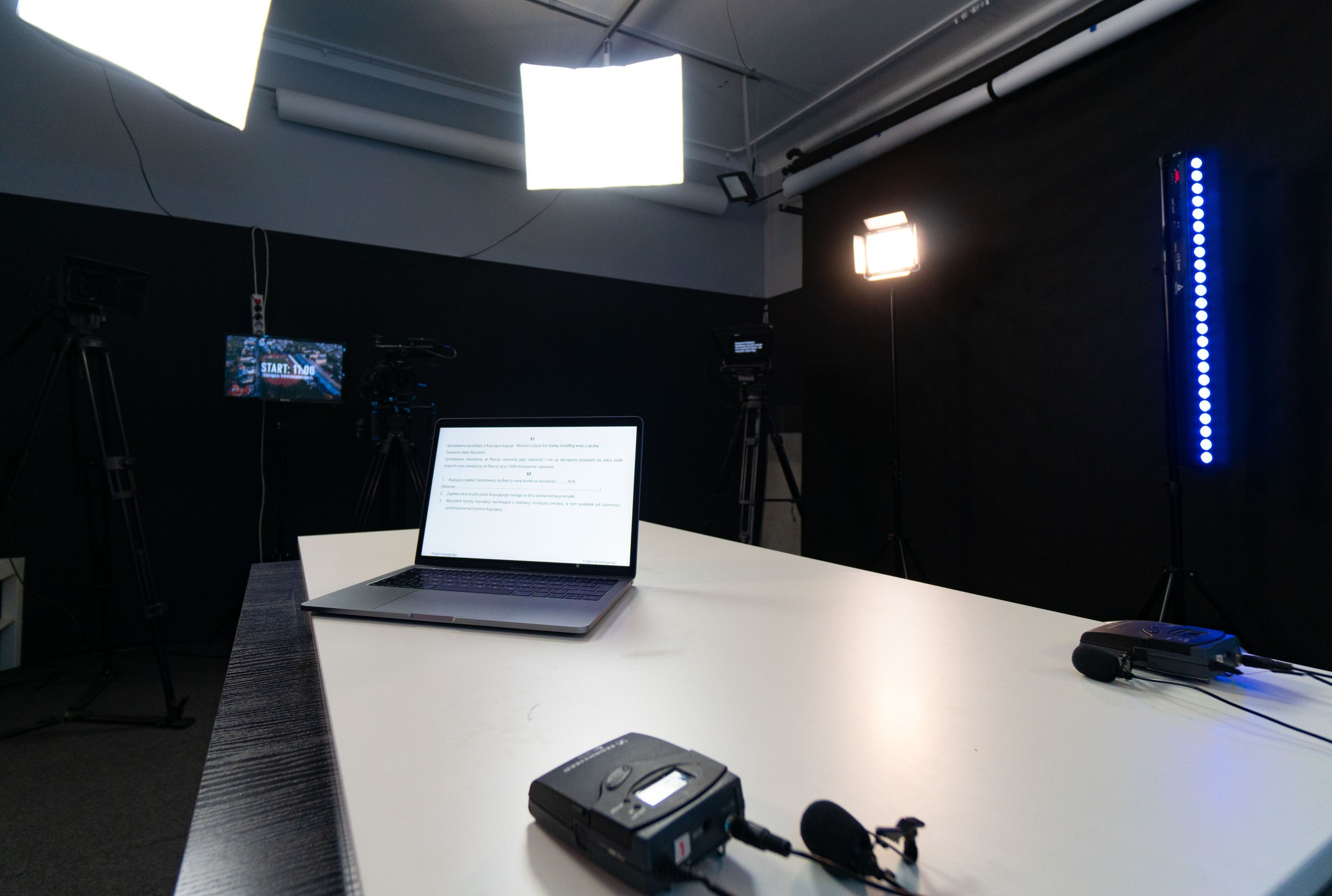 Wyposażone studio nagraniowe z oświetleniem studyjnym, mikrofonem krawatowym, laptopem z widocznym tekstem umowy sprzedaży sprzętu i kamerami na statywach w tle.