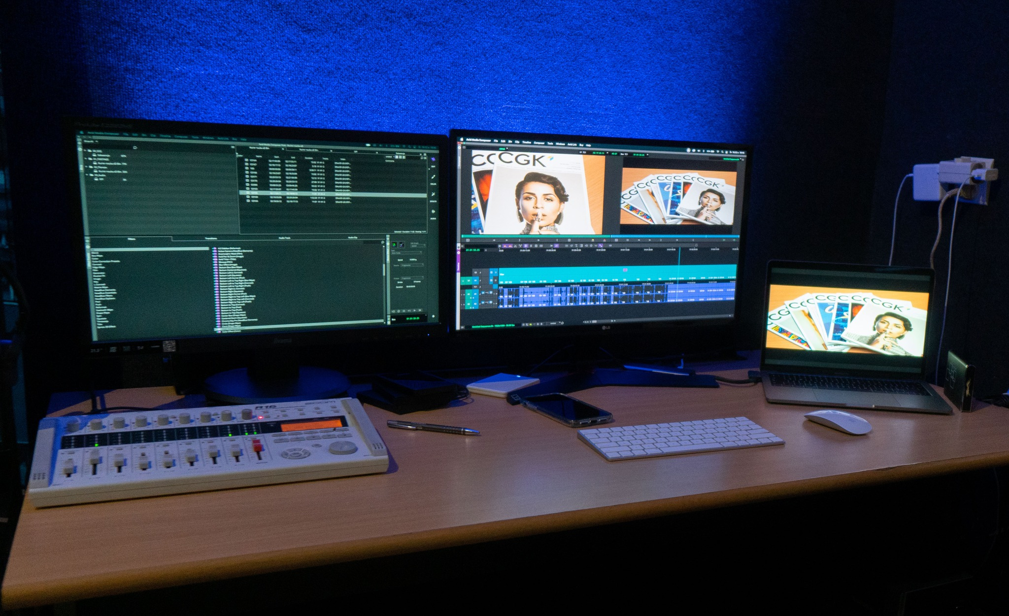 Stanowisko do postprodukcji wideo z dwoma monitorami wyświetlającymi timeline projektu, laptopem z otwartym projektem, mikserem audio i akcesoriami na biurku, oświetlone niebieskim światłem.