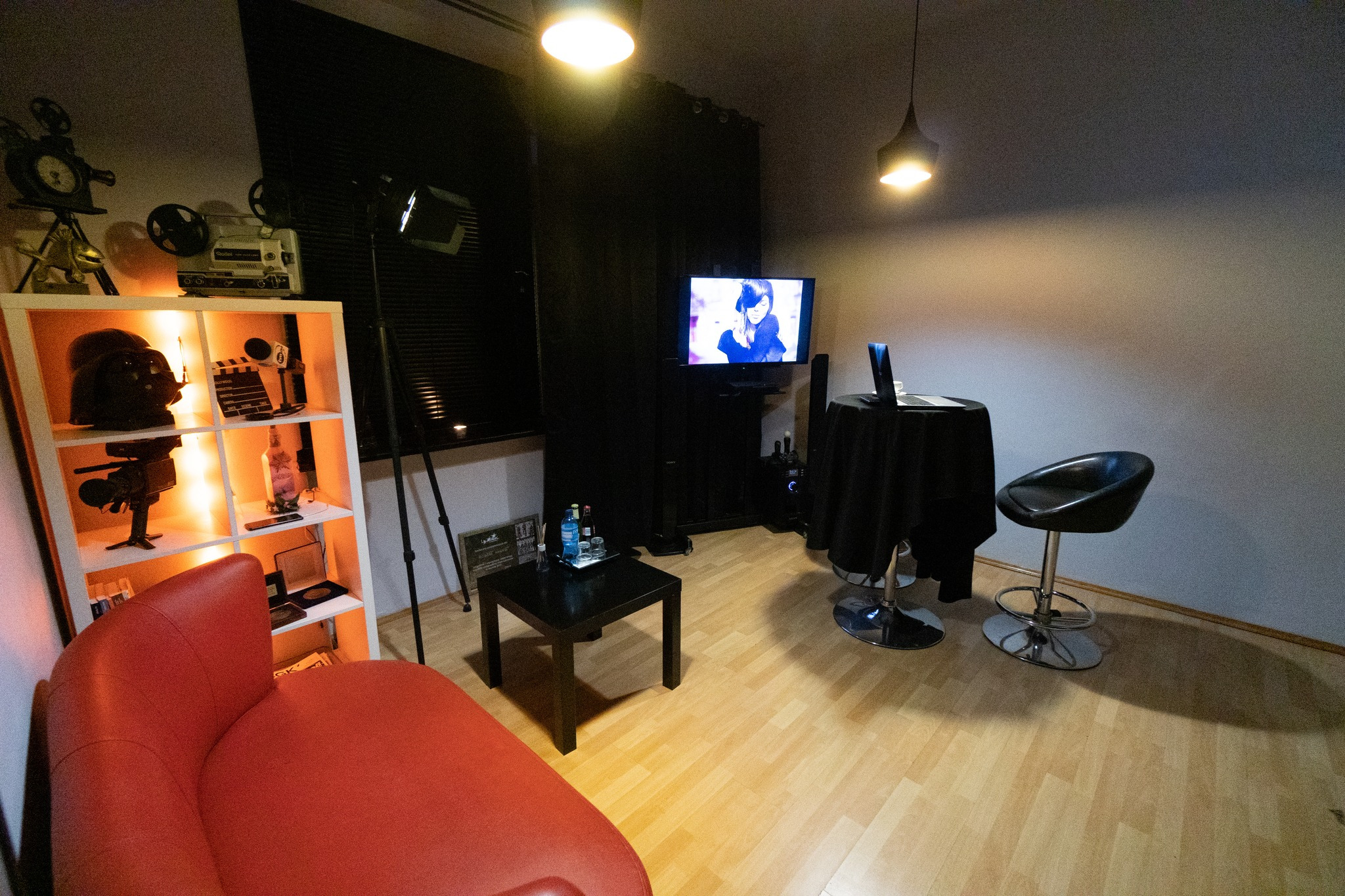 Wnętrze studia filmowego z rekwizytami: stara kamera, klaps filmowy, projektor na półce z podświetleniem, monitor z widoczną sceną filmową, stolik z napojami, czerwony fotel i czarne zasłony.