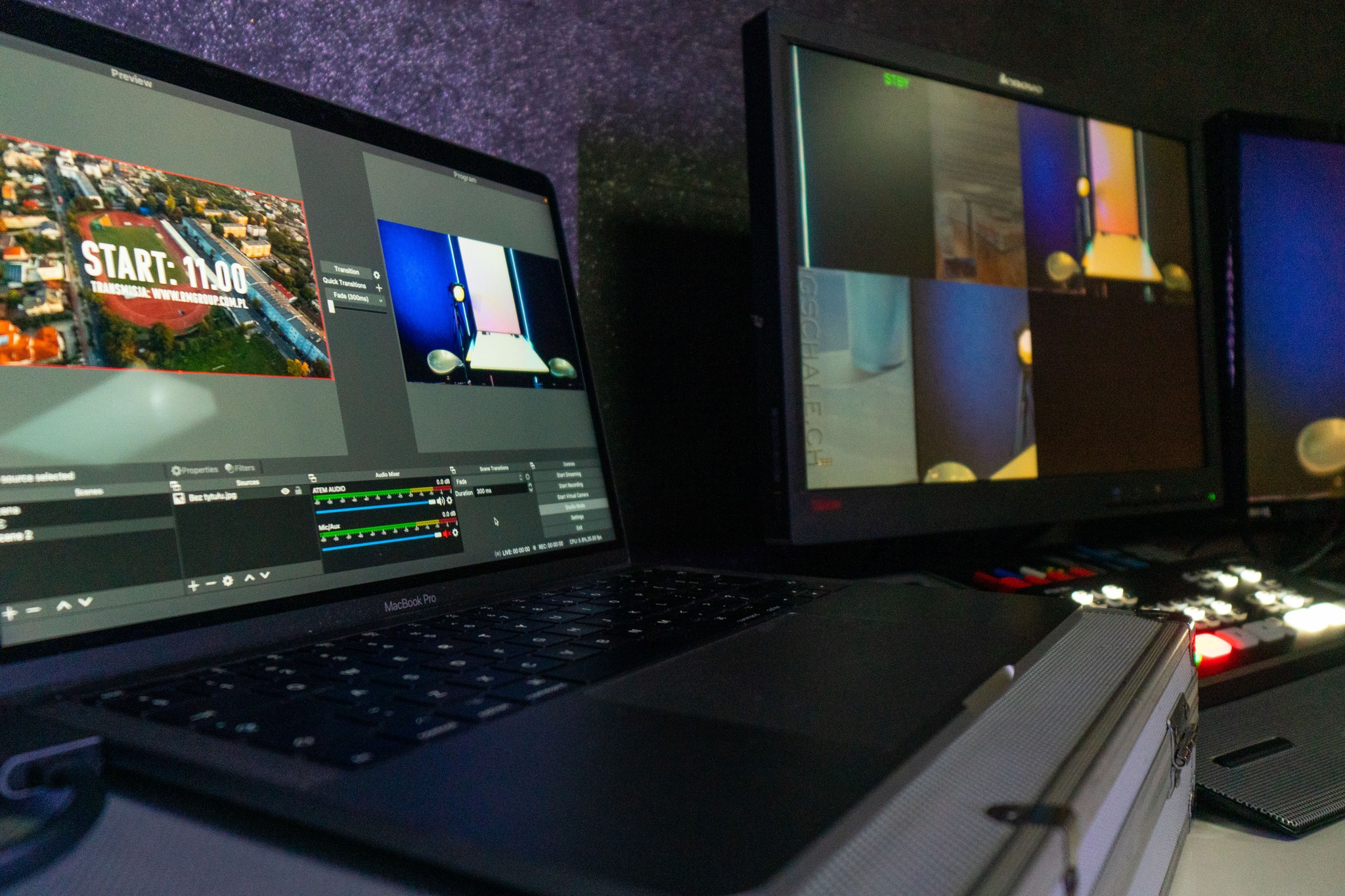 Stanowisko do streamingu z laptopem MacBook Pro, mikserem audio ATEM i monitorami podglądowymi. Na ekranie laptopa widoczny program do streamingu z podglądem transmisji 'START: 11:00'.