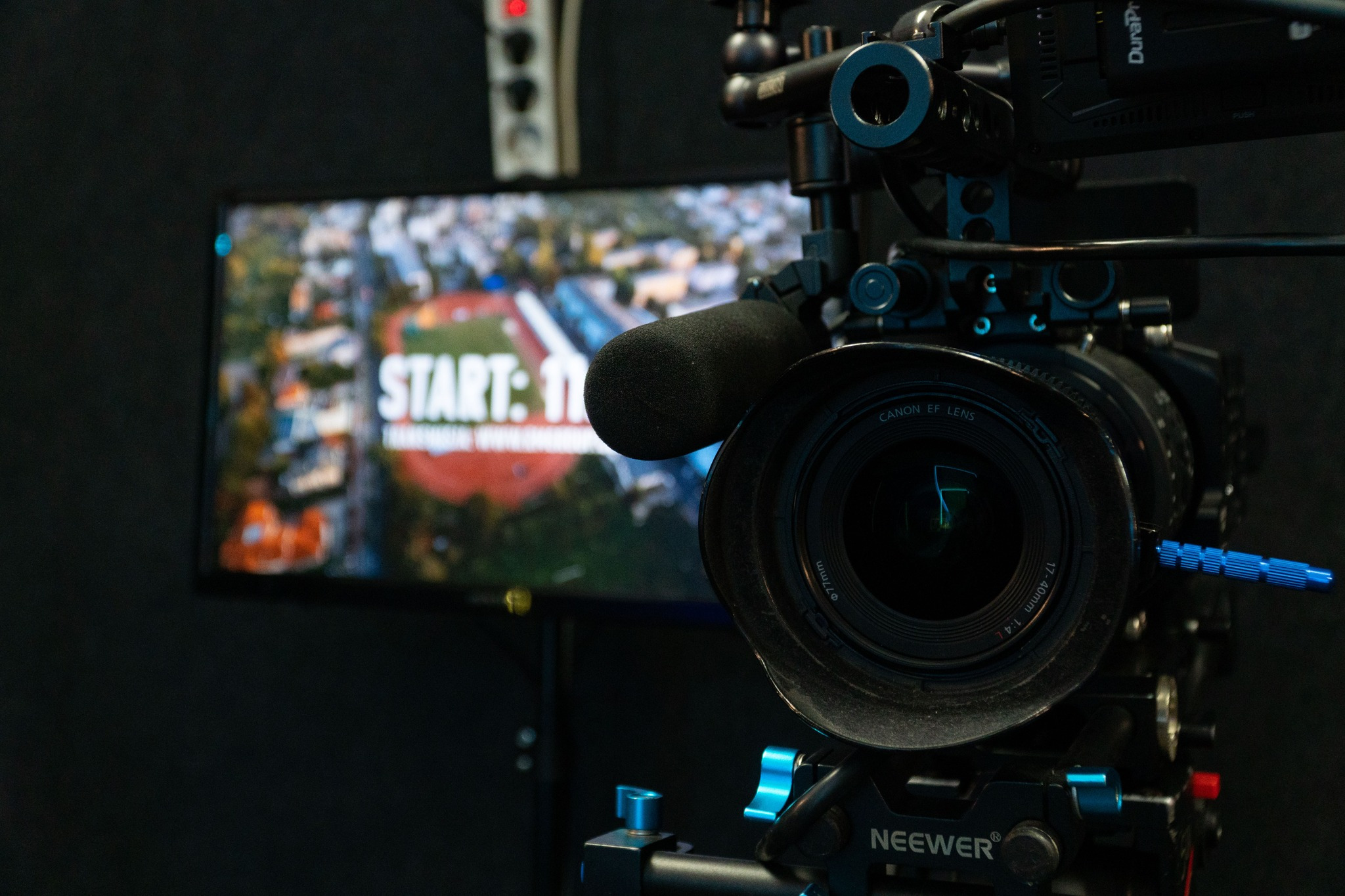 Profesjonalna kamera z mikrofonem na statywie, obiektyw Canon EF 17-40mm, w tle monitor z wyświetlanym obrazem z nagrania przedstawiającego stadion z napisem 'START: 11'.