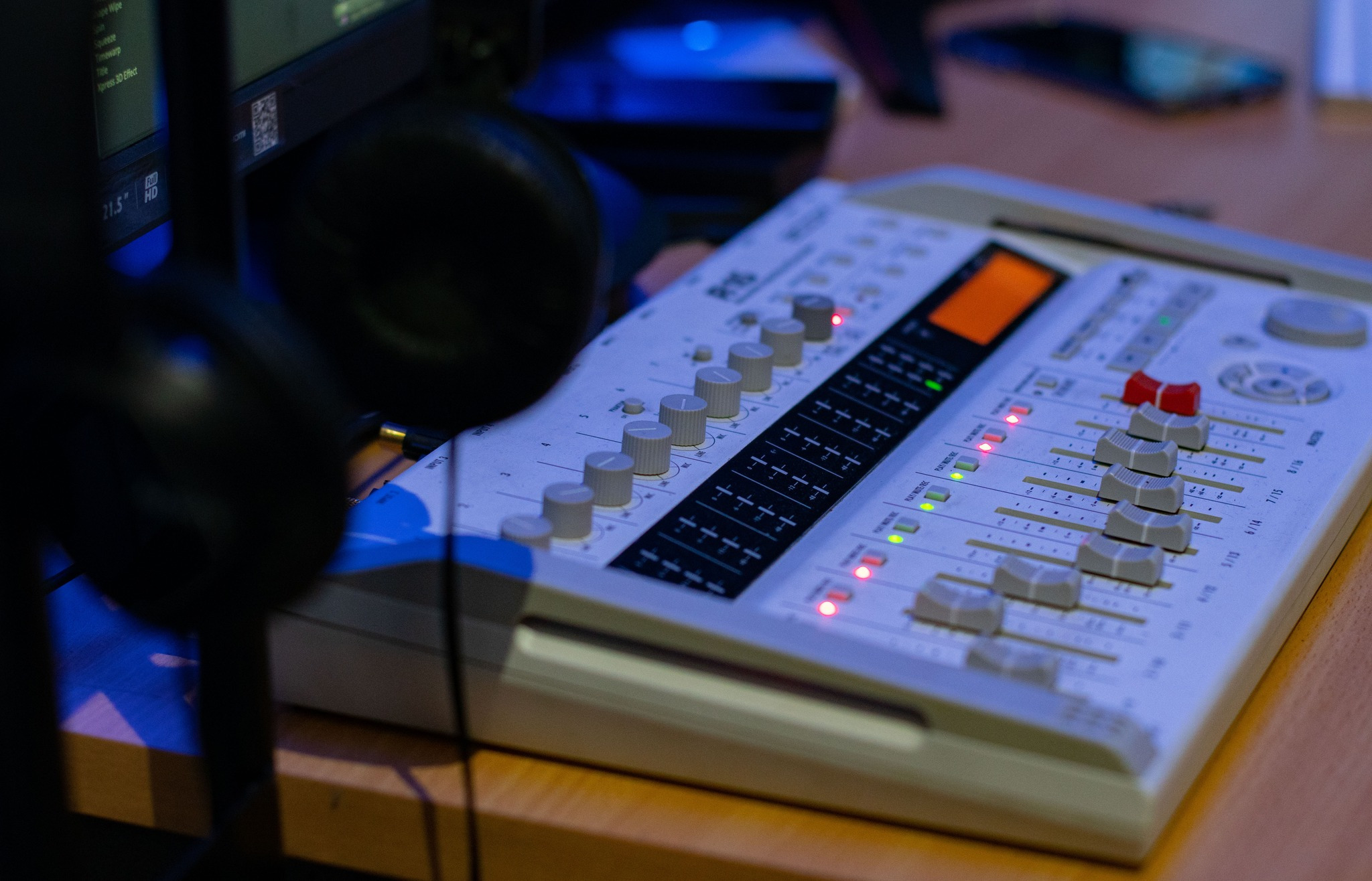 Mikser audio na drewnianym biurku, z widocznymi suwakami, pokrętłami i podświetlonymi diodami. W tle słuchawki i monitor z interfejsem programu do edycji wideo.