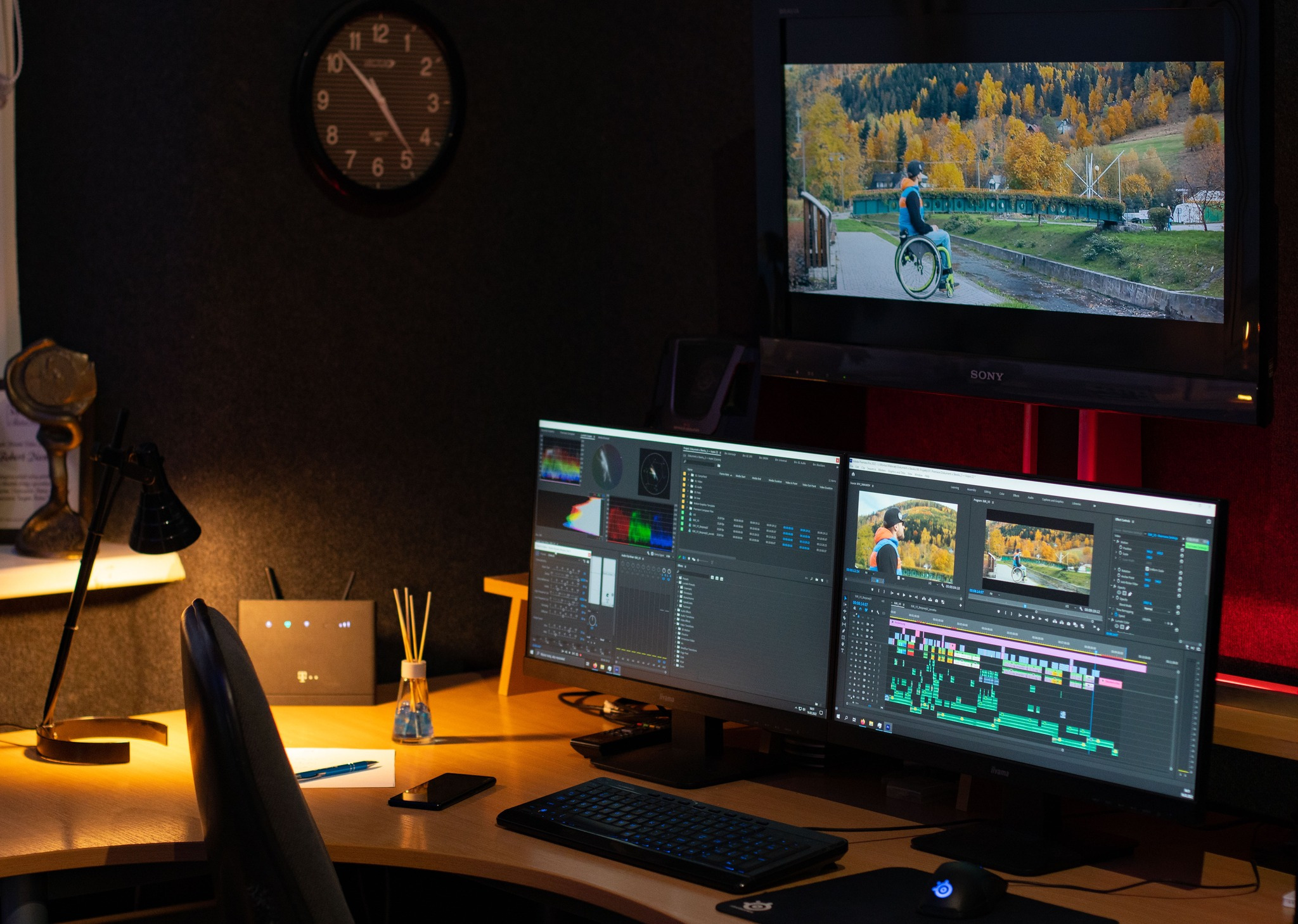 Stanowisko montażowe z dwoma monitorami, na których widoczny jest interfejs programu do montażu wideo z osią czasu i podglądem ujęć jesiennego krajobrazu; nad nimi telewizor Sony wyświetlający...