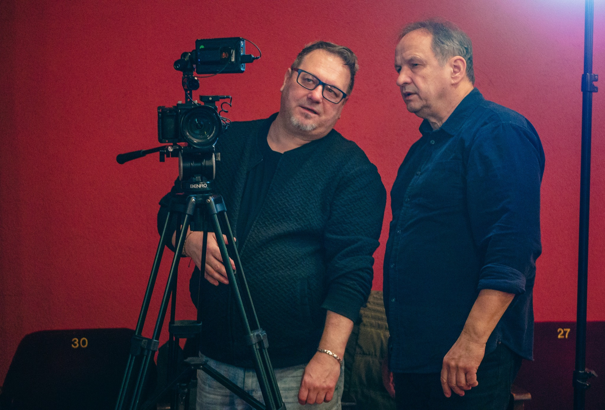 Dwaj mężczyźni przy kamerze Benro na statywie, podczas kręcenia ujęcia na tle czerwonej ściany z numerami siedzeń 30 i 27.