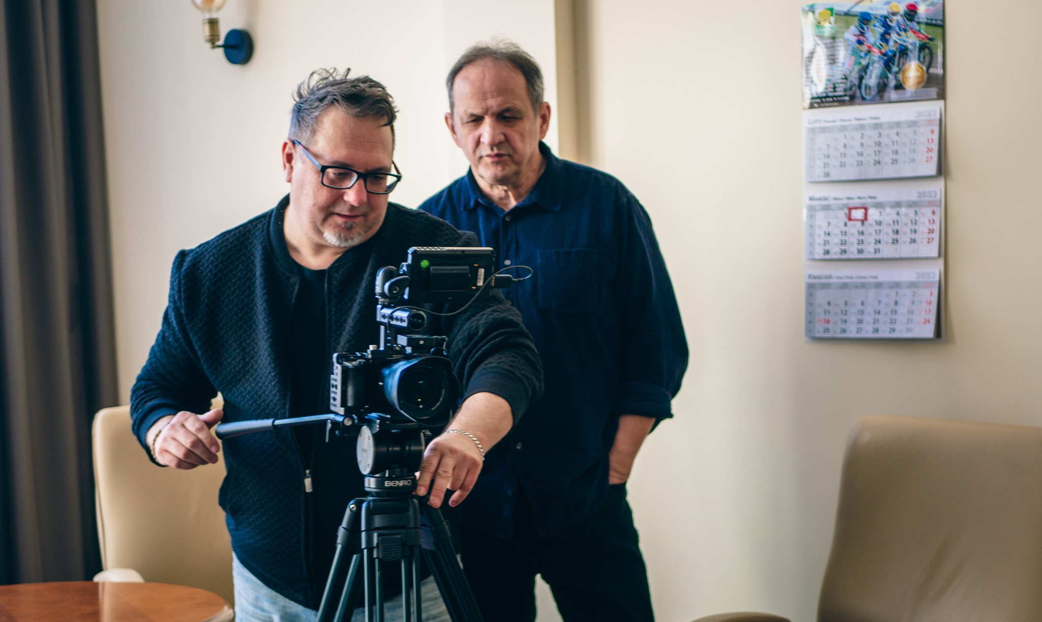 Operator kamery w okularach reguluje ustawienia kamery Benro na statywie podczas kręcenia filmu dokumentalnego, drugi mężczyzna obserwuje. Na ścianie wisi kalendarz.