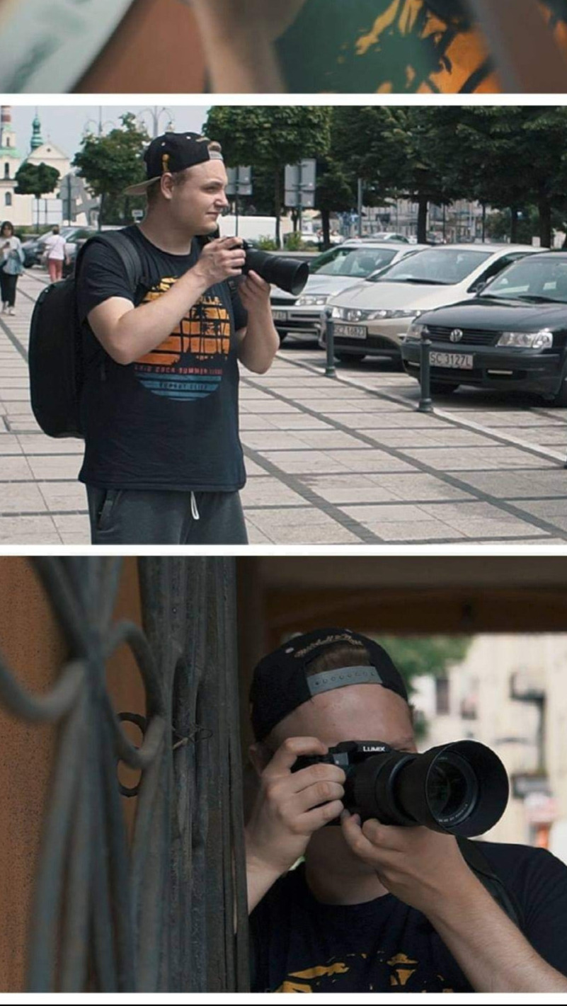 Mężczyzna w czarnej koszulce z aparatem fotograficznym w ręku, stojący na tle ulicy z zaparkowanymi samochodami i kościołem w oddali; ujęcie zza metalowej bramy.