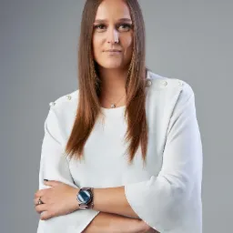 Portret kobiety w białej bluzce z rękawami typu dzwon, z rękami skrzyżowanymi, zegarkiem na nadgarstku i delikatną biżuterią, na jednolitym szarym tle.