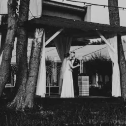 Czarno-białe zdjęcie pary młodej w objęciach pod drewnianą altaną z białymi zasłonami, drzewa wkomponowane w konstrukcję, w tle budynek z oknami.