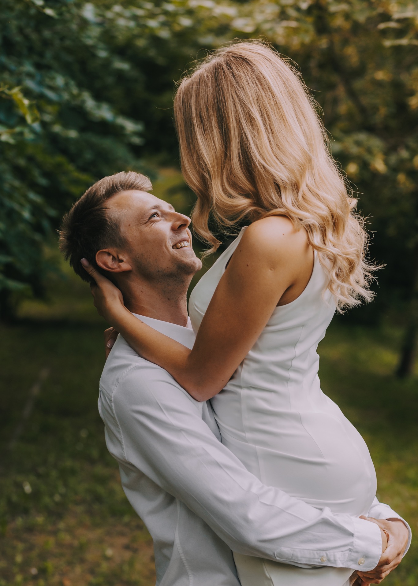 Uśmiechnięty mężczyzna w białej koszuli trzyma w ramionach kobietę w białej sukience ciążowej, patrząc na nią z czułością, w plenerze w Sokółce.