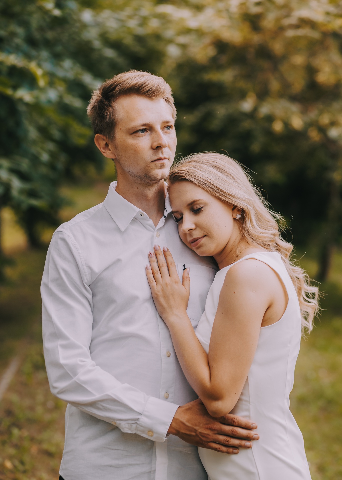 Para w jasnych ubraniach przytulona na tle zieleni, kobieta z blond włosami opiera głowę o ramię mężczyzny, subtelny uśmiech.