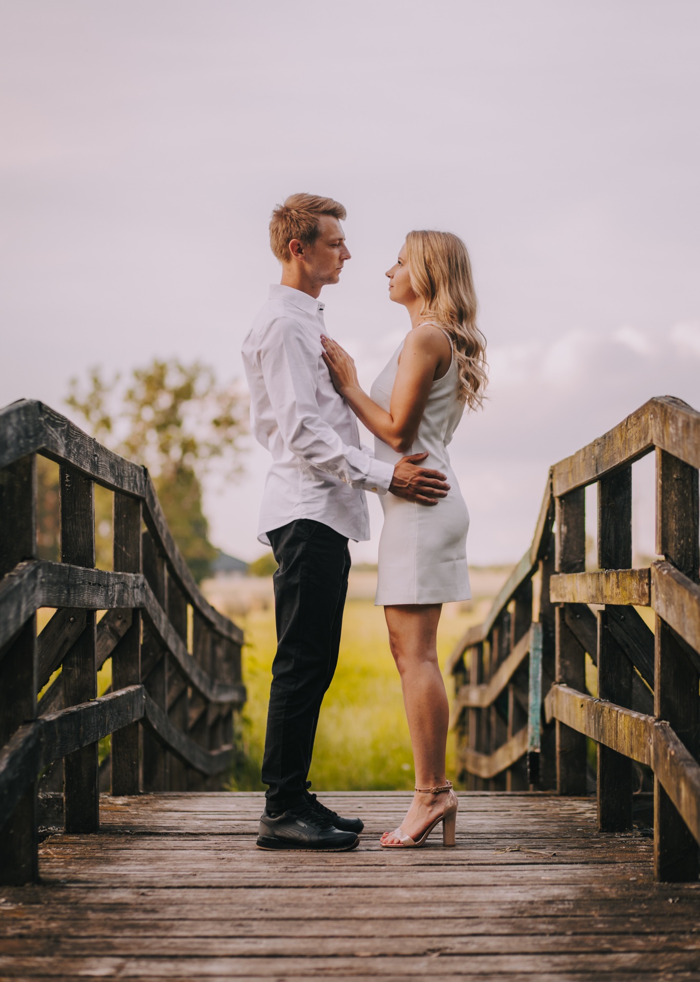 Para zakochanych na drewnianym moście, mężczyzna w białej koszuli i czarnych spodniach, kobieta w białej sukience i beżowych szpilkach, delikatny dotyk dłoni na piersi, spojrzenia pełne uczucia,...
