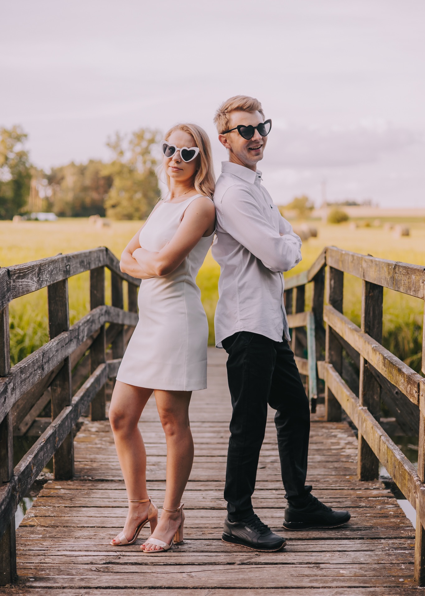 Para młoda w okularach w kształcie serc, oparta plecami o siebie na drewnianym mostku z widokiem na pole ze słomianymi balotami w tle.