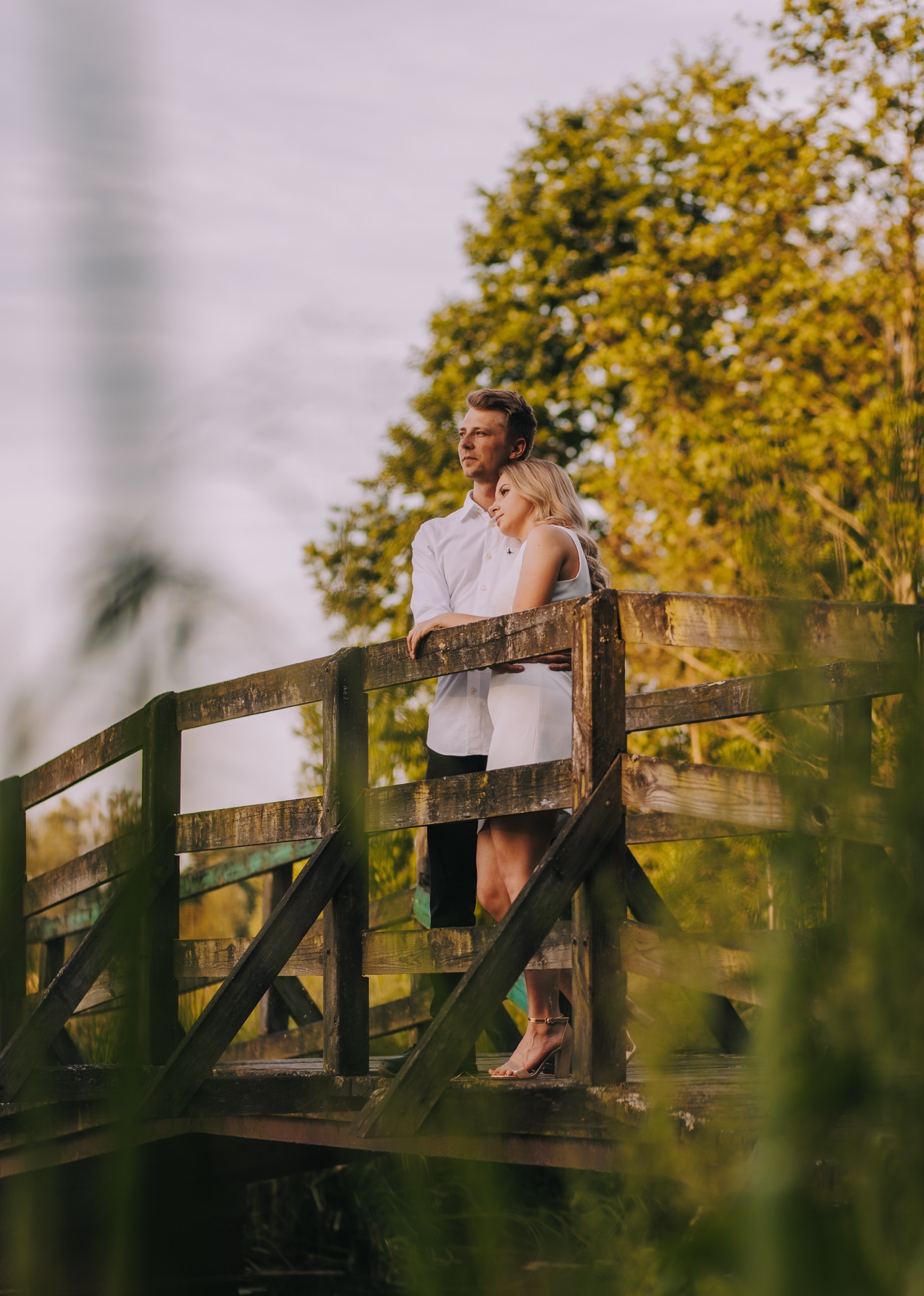Para zakochanych, on w białej koszuli i czarnych spodniach, ona w białej sukience i złotych sandałach na obcasie, stoi na drewnianym mostku, spoglądając w dal podczas sesji ślubnej w plenerze.