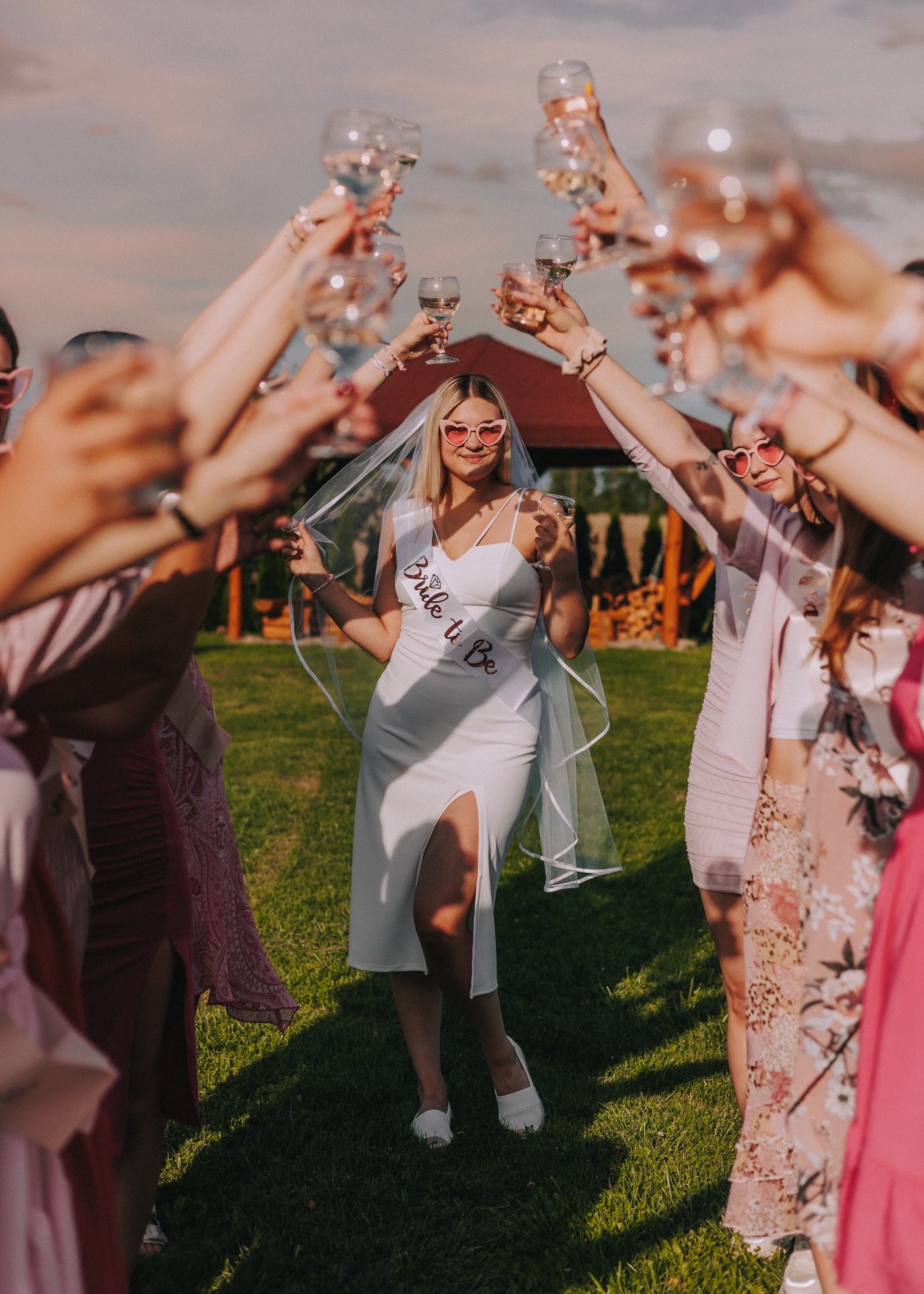 Panna młoda w białej sukience i welonie z napisem 'Bride to Be' w otoczeniu przyjaciółek w różowych sukienkach wznosi toast w Sokółce. Wszystkie kobiety mają na sobie różowe okulary w kształcie serc.