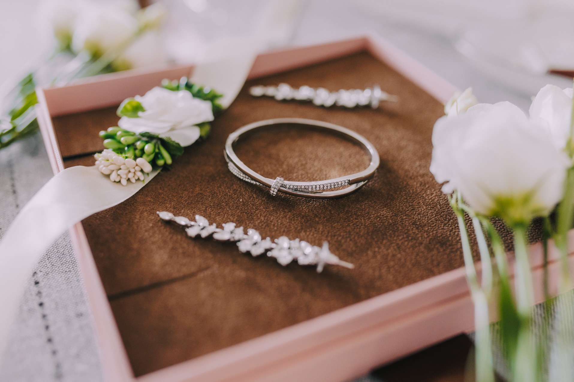 Srebrna bransoletka z cyrkoniami, spinka do włosów z motywem liści, butonierka z białych róż i wstążką, umieszczone w różowym pudełku na brązowym tle, elementy dekoracyjne ślubne.