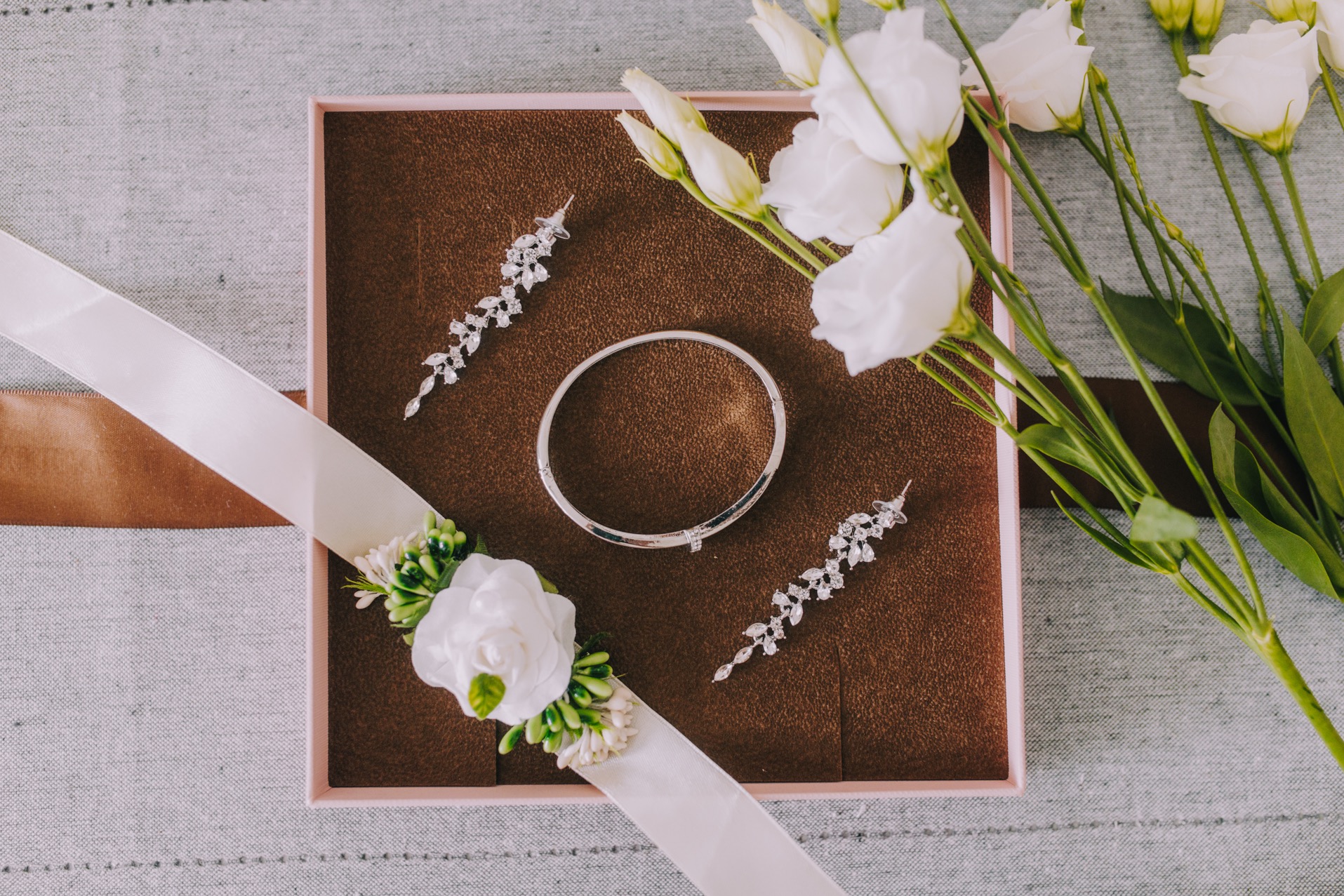 Srebrna bransoletka, kolczyki z kryształkami i butonierka z białych róż na satynowej wstążce w pudełku, obok bukiet białych eustom.