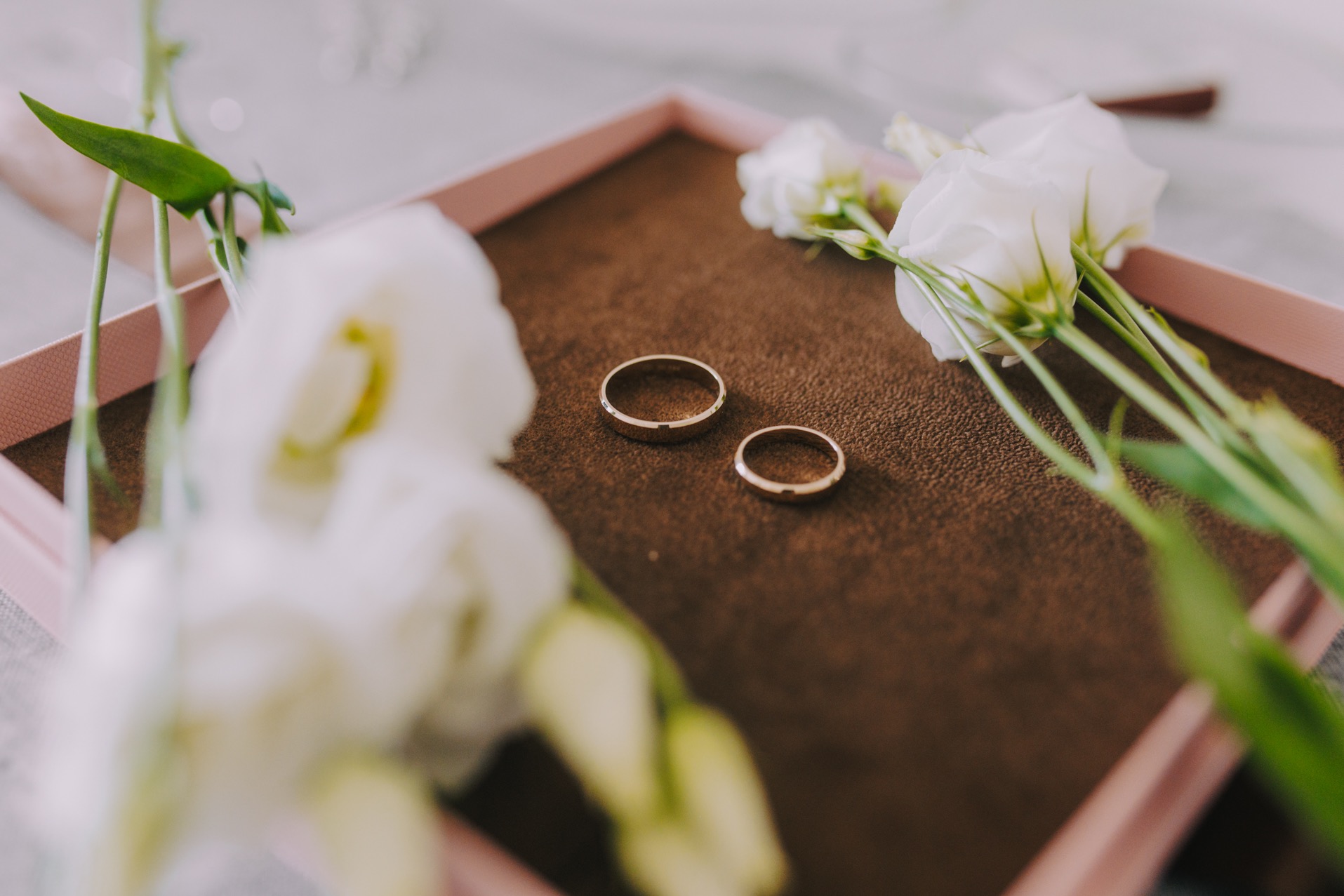 Dwa złote obrączki ślubne ułożone na brązowym tle w różowym pudełku, ozdobione białymi kwiatami eustomy.